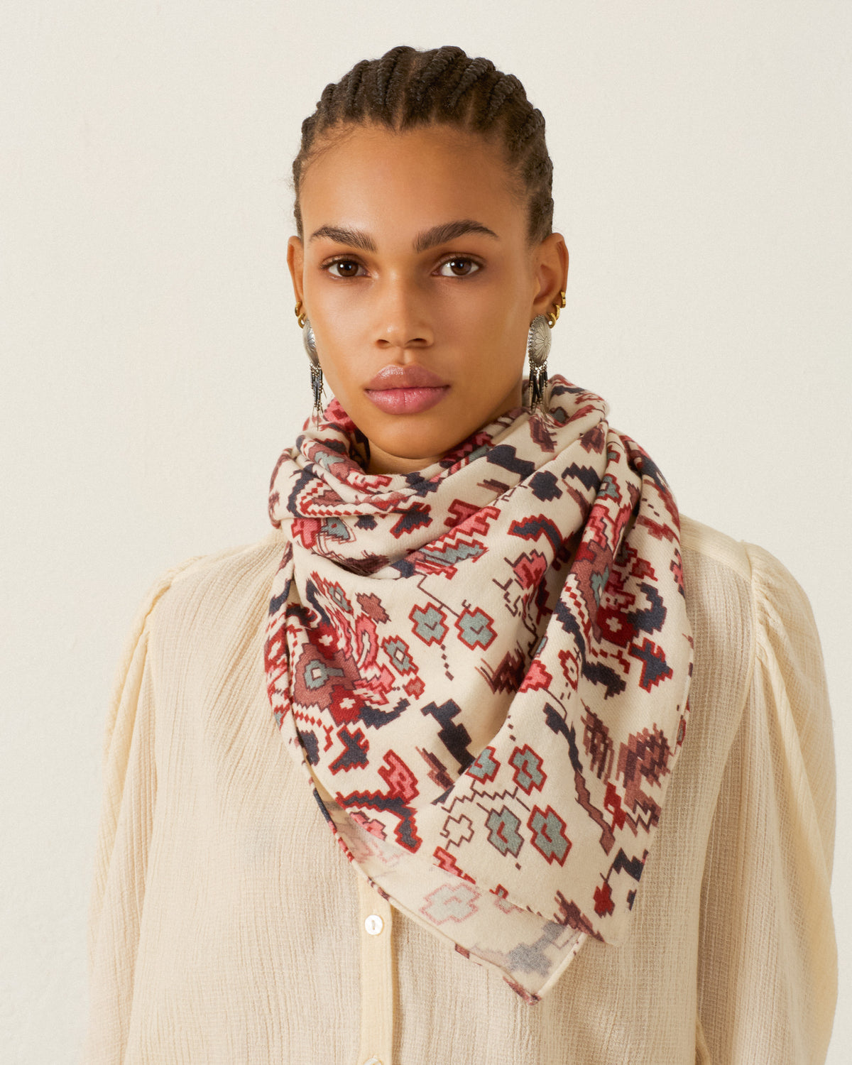 A woman with braided hair wears a cream blouse and the Fable Horizon Kilim Scarf—an accessory in red, blue, and beige tones with hand-finished edges—as she stands against a plain light background.