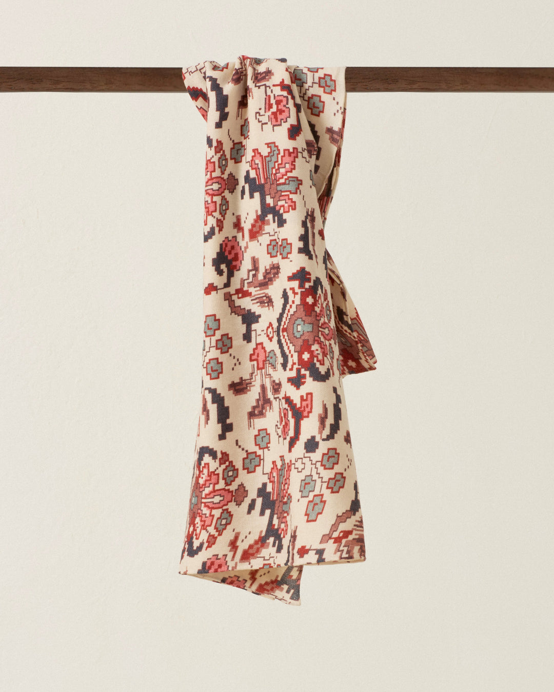 The Fable Horizon Kilim Scarf, crafted from beige fabric with a red, blue, and black abstract pattern, is draped over a dark wooden rod against a plain light background. Inspired by traditional kilim designs, this scarf features hand-finished edges for an artisanal touch.