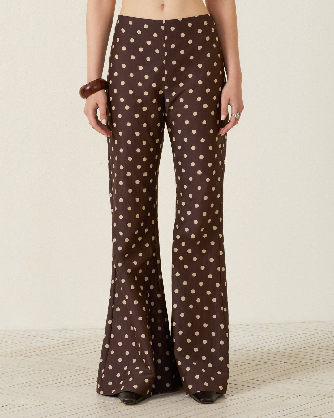 A person shown from the waist down wears the Farah Mahogany Dot Pant—high-waisted, brown flared trousers with beige polka dots, made from a soft cotton-silk blend—standing against a plain background.