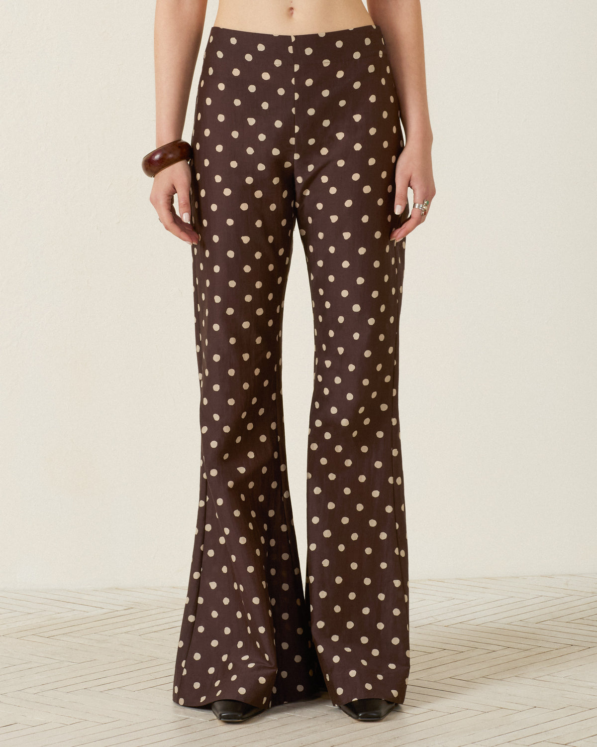 A person shown from the waist down wears the Farah Mahogany Dot Pant—high-waisted, brown flared trousers with beige polka dots, made from a soft cotton-silk blend—standing against a plain background.