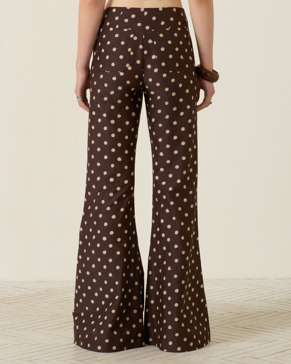 A person stands facing away, wearing the Farah Mahogany Dot Pant—a high-waisted trouser crafted from a luxurious cotton-silk blend, featuring a beige polka dot pattern and a stylish bell-bottom flare.