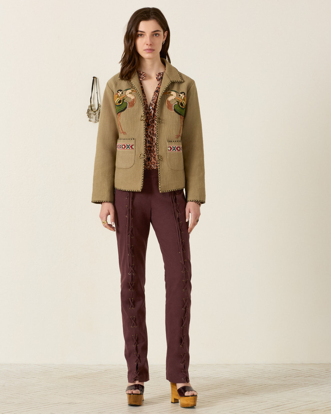 A woman stands facing forward, wearing a beige jacket with bird embroidery, a patterned blouse, and the Razza Mahogany Pant—cotton straight-leg pants featuring grommeted lace-up ties. She pairs the look with platform heels, while a small purse hangs on the wall behind her.