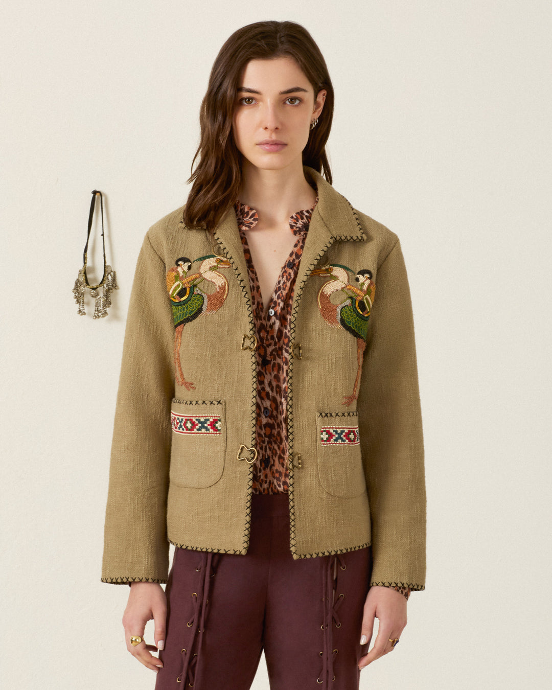 A woman stands against a plain wall, wearing the Francis Heron Rider Jacket over a patterned blouse and maroon pants with lace detailing; jewelry hangs on the wall beside her.