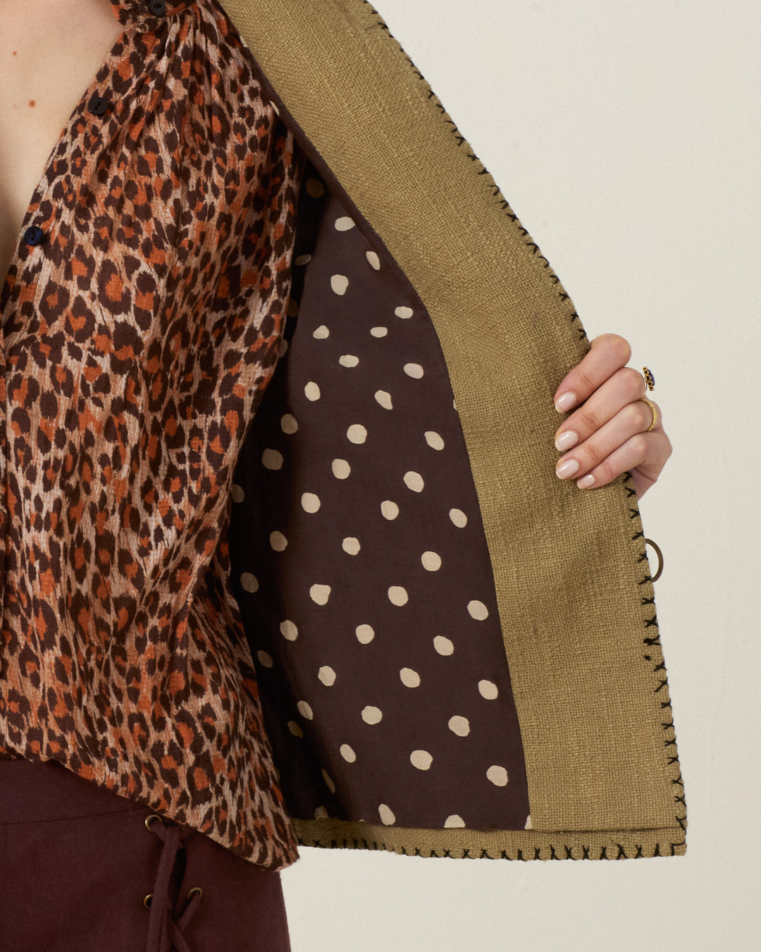 A person holds open the Francis Heron Rider Jacket, made of tan khadi cotton and lined with brown fabric featuring large white polka dots, revealing a leopard print blouse underneath.