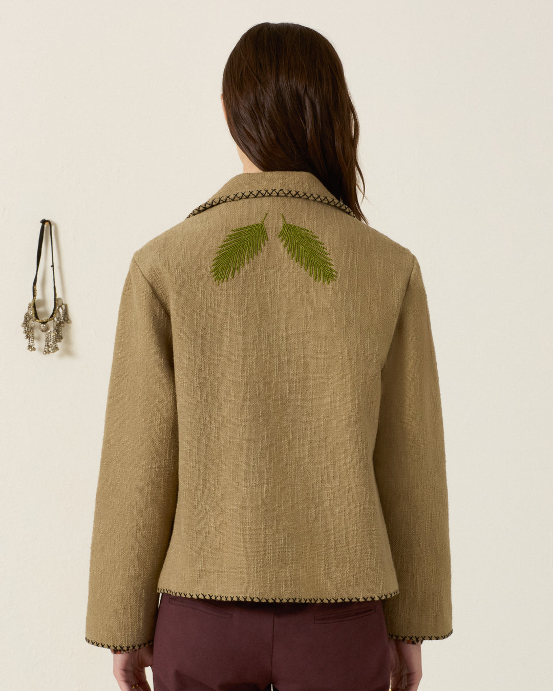 A person with long brown hair wears the Francis Heron Rider Jacket, a tan piece featuring green leaf embroidery on the back, while standing against a plain wall adorned with a hanging necklace.