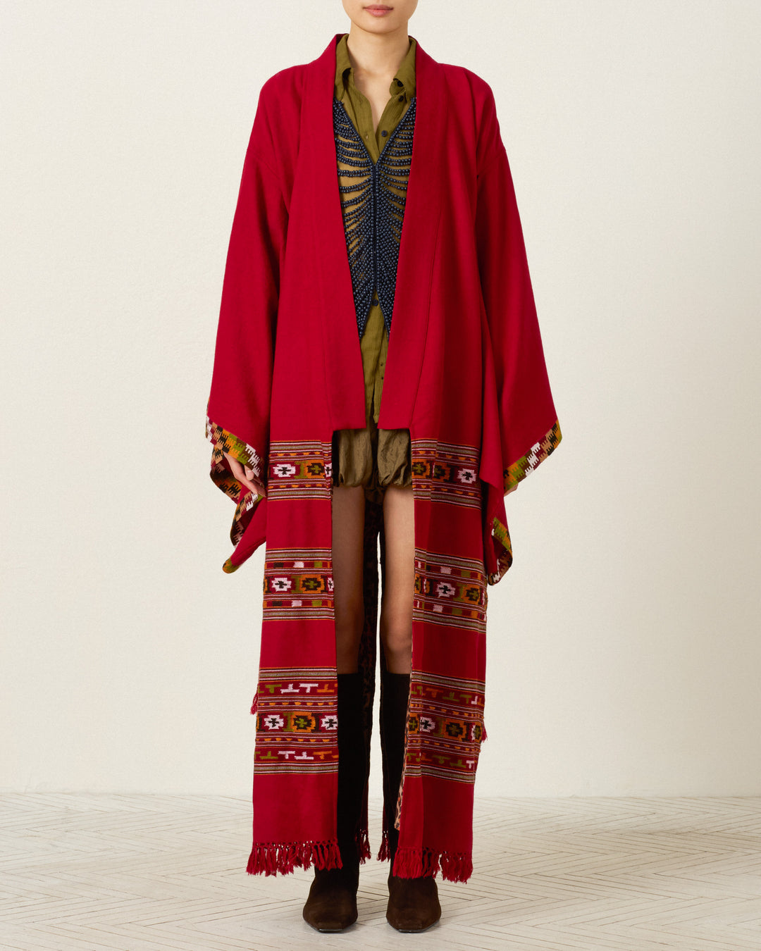 A person wearing the Grace Chili Tree Wool Coat—a long red patterned outerwear piece inspired by Himalayan artisan garments—layered over a green shirt, shorts, and black knee-high boots, standing against a plain background.
