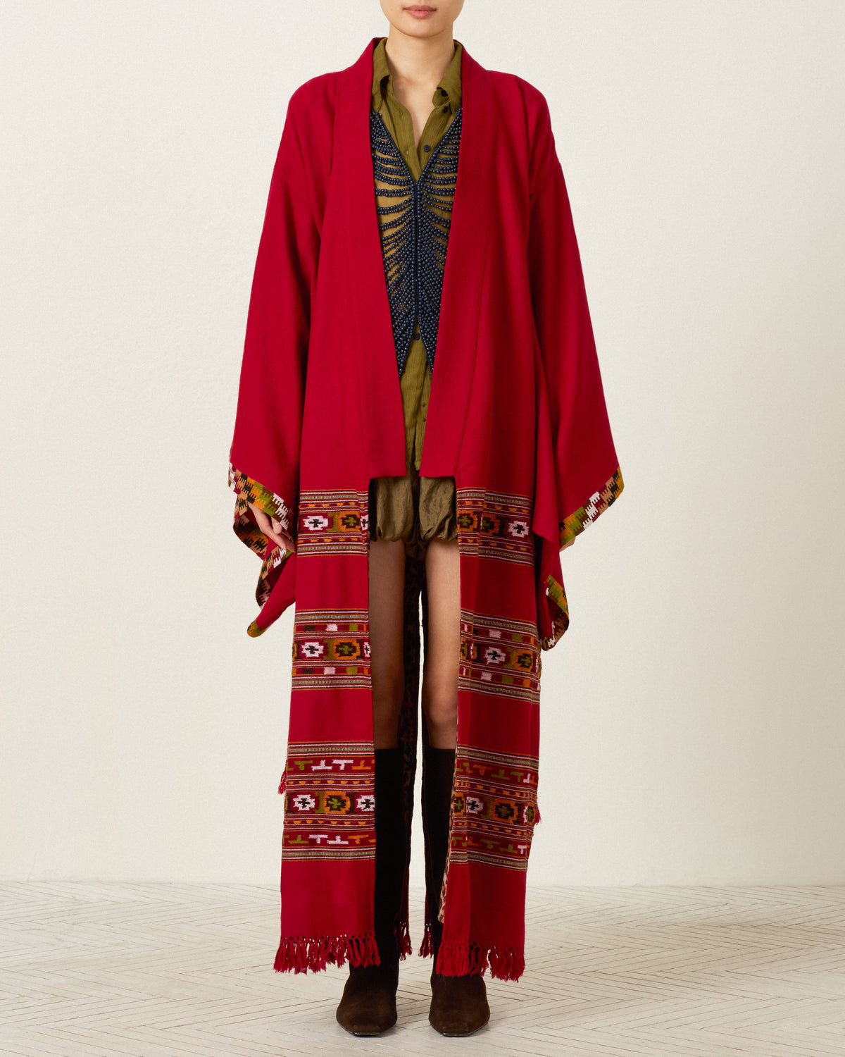 A person wearing the Grace Chili Tree Wool Coat—a long red patterned outerwear piece inspired by Himalayan artisan garments—layered over a green shirt, shorts, and black knee-high boots, standing against a plain background.