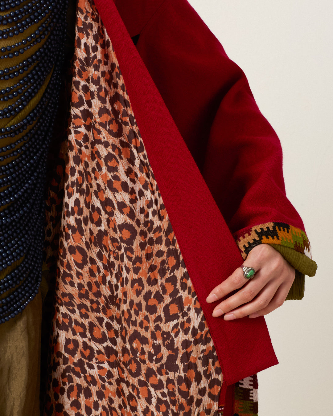 A person holds open the Grace Chili Tree Wool Coat—a red Himalayan artisan wool belted wrap coat with a leopard print lining, displaying its green and brown patterned cuffs while wearing a turquoise ring.