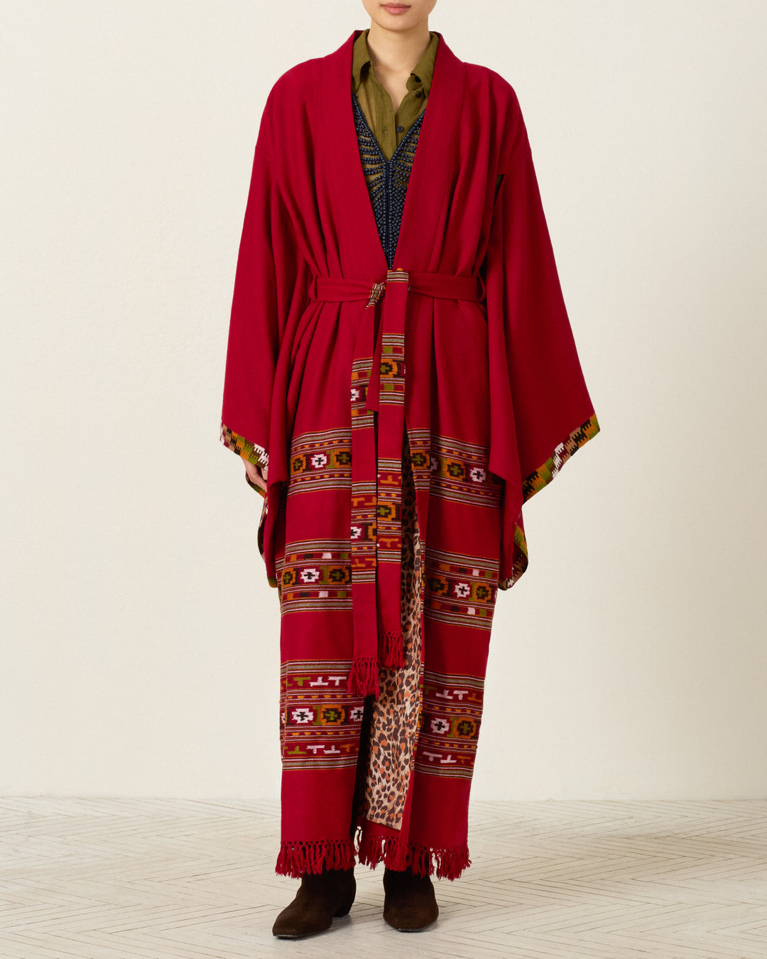 A person stands against a plain background, wearing the Grace Chili Tree Wool Coat—a long red robe-style coat inspired by Himalayan artisan garments, featuring colorful patterned stripes along the lower half, a belted waist, and paired with brown boots.