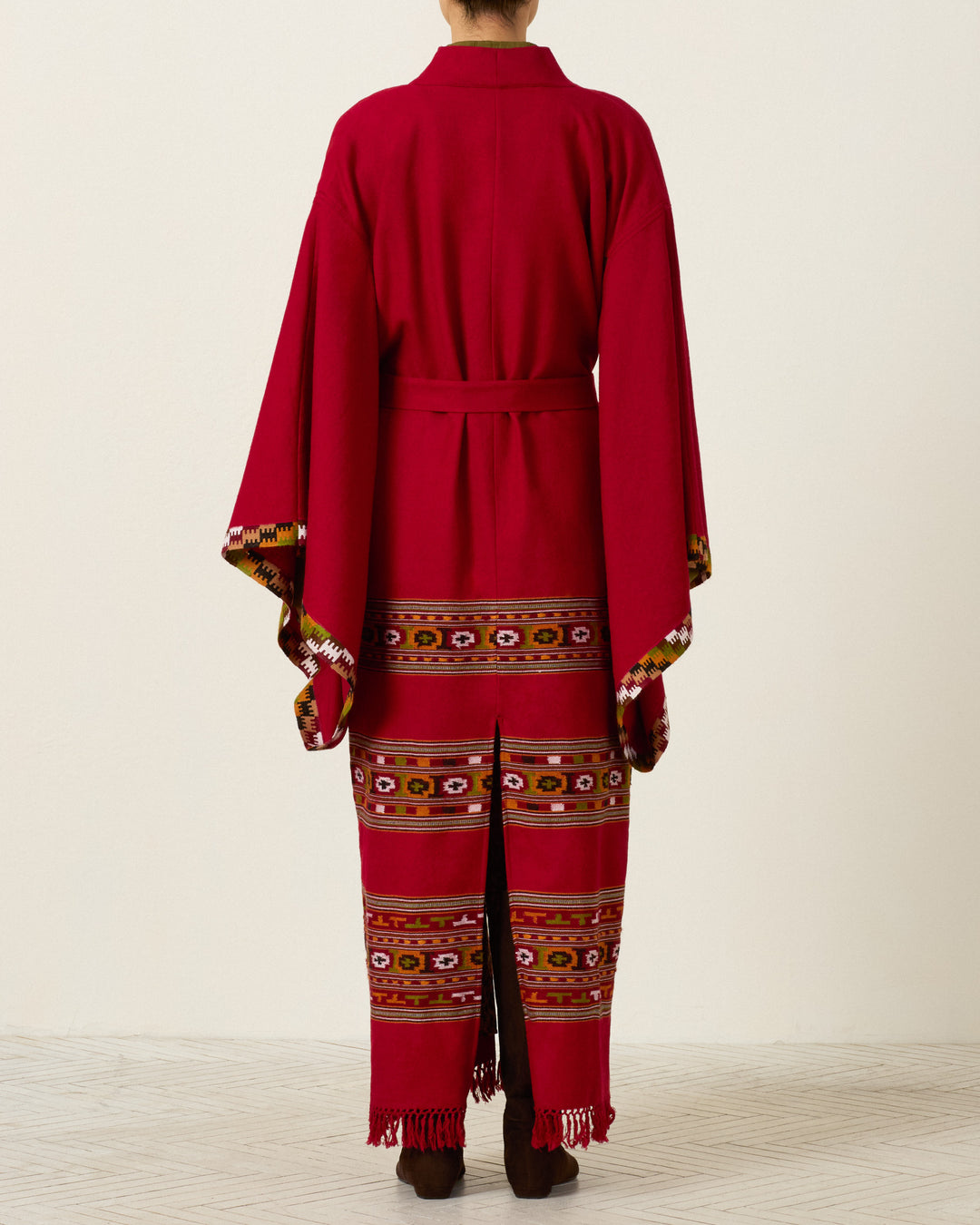 A person stands with their back to the camera, wearing the Grace Chili Tree Wool Coat—a long red robe inspired by Himalayan artisan garments, featuring colorful geometric patterns and fringed hems—set against a plain light background.
