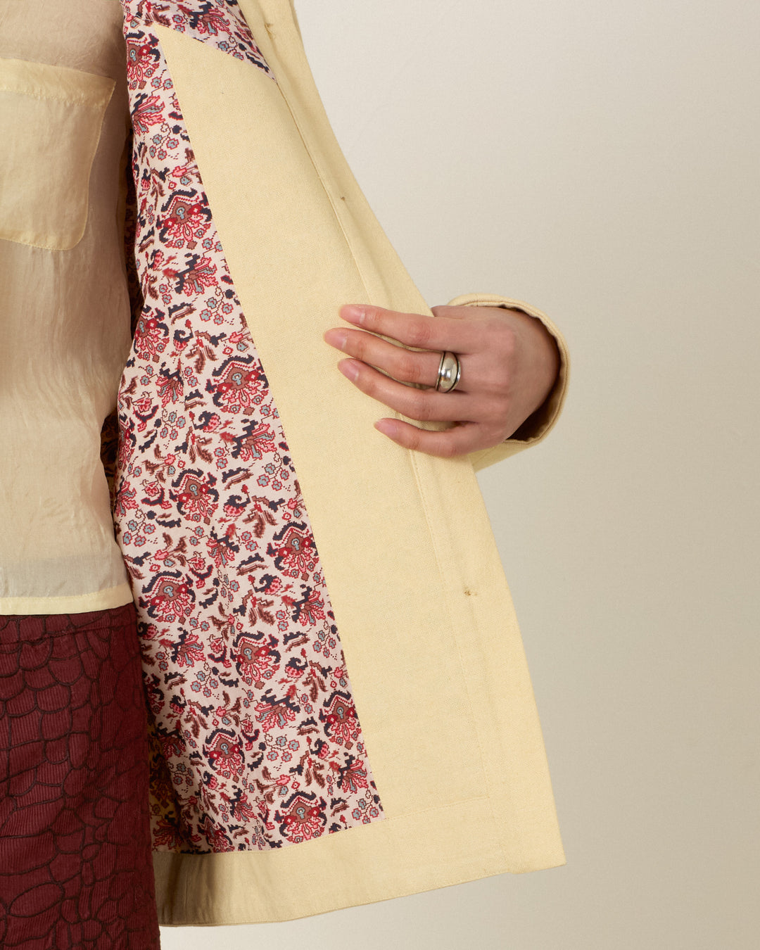 Wearing the Judson Jungle Book Jacket, a person holds open its safari-inspired light yellow outer layer to reveal a vibrant red and white floral patterned lining, paired with a sheer shirt and maroon skirt.