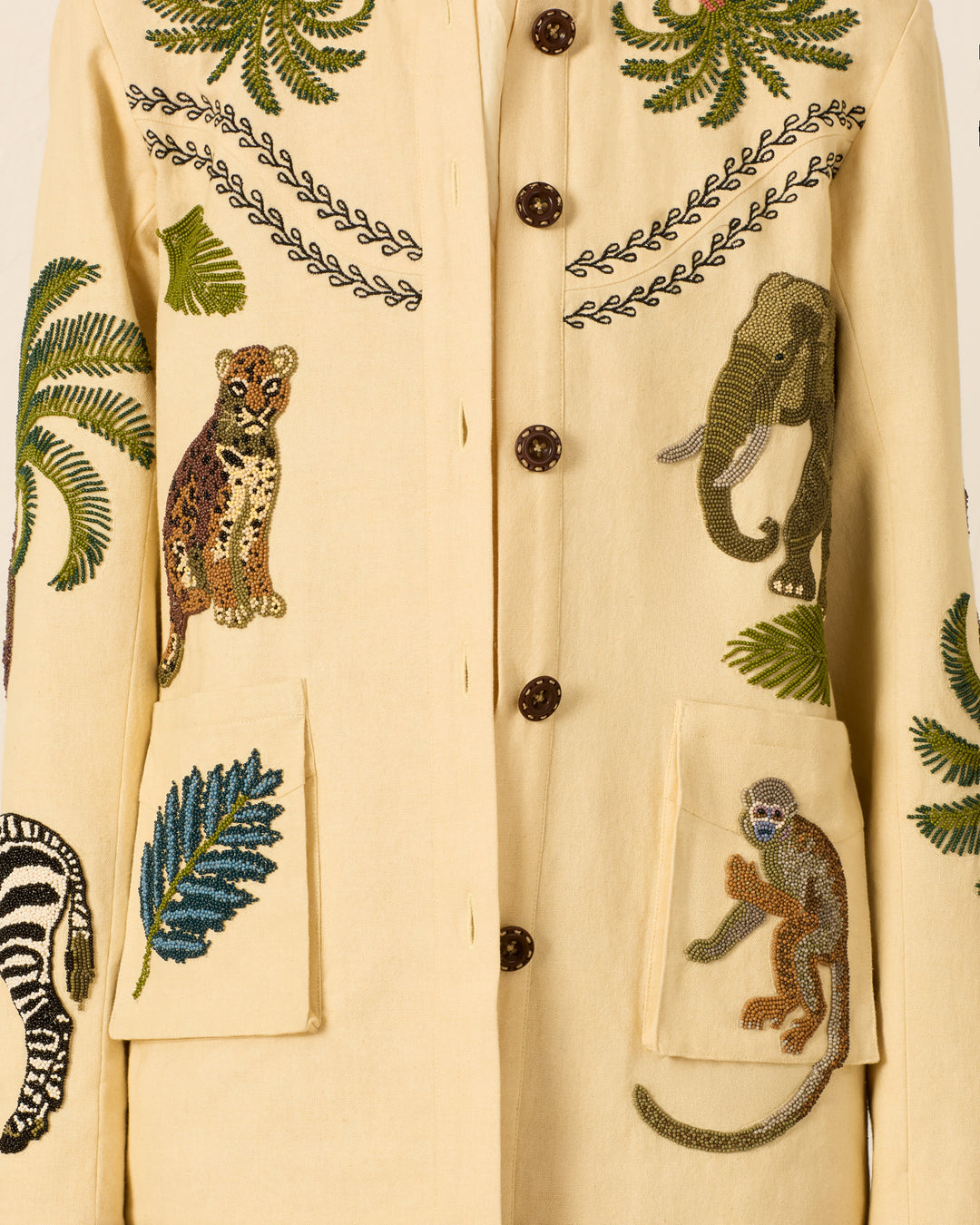 Introducing the Judson Jungle Book Jacket—a beige jacket adorned with brown buttons and intricate embroidery featuring a cheetah, elephant, zebra, monkey, and vibrant green leaves on the front and sleeves. This safari-inspired statement piece infuses any wardrobe with adventurous charm and playful detail.