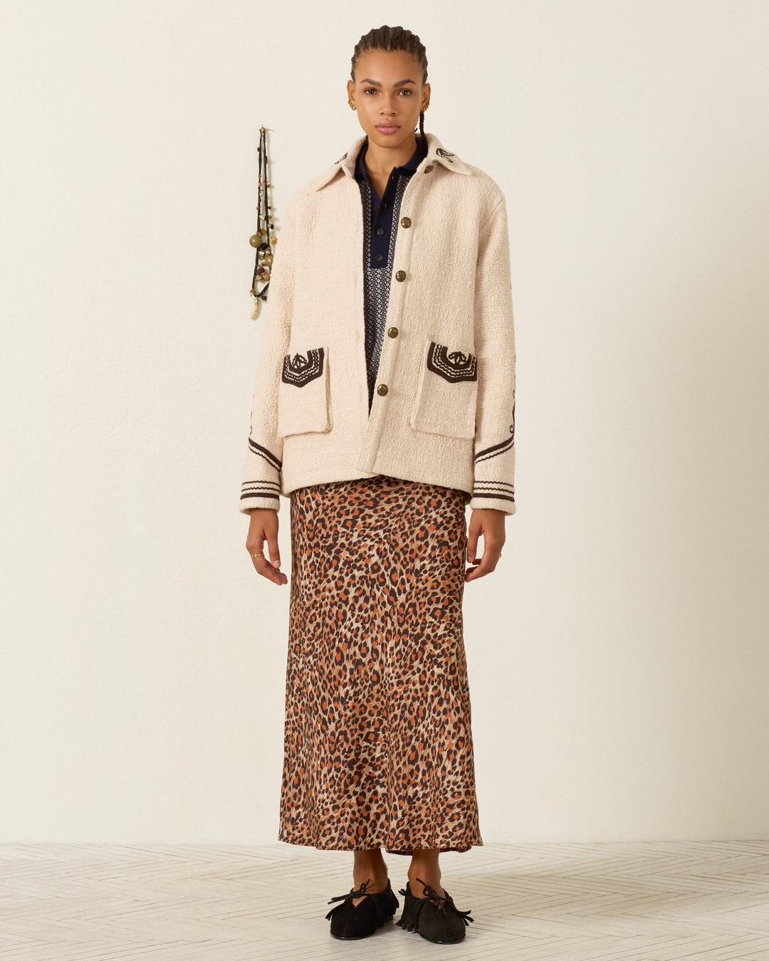 A person stands indoors against a plain wall, wearing the Jude Bushland Cord Jacket, styled with a navy shirt and a long leopard print skirt.