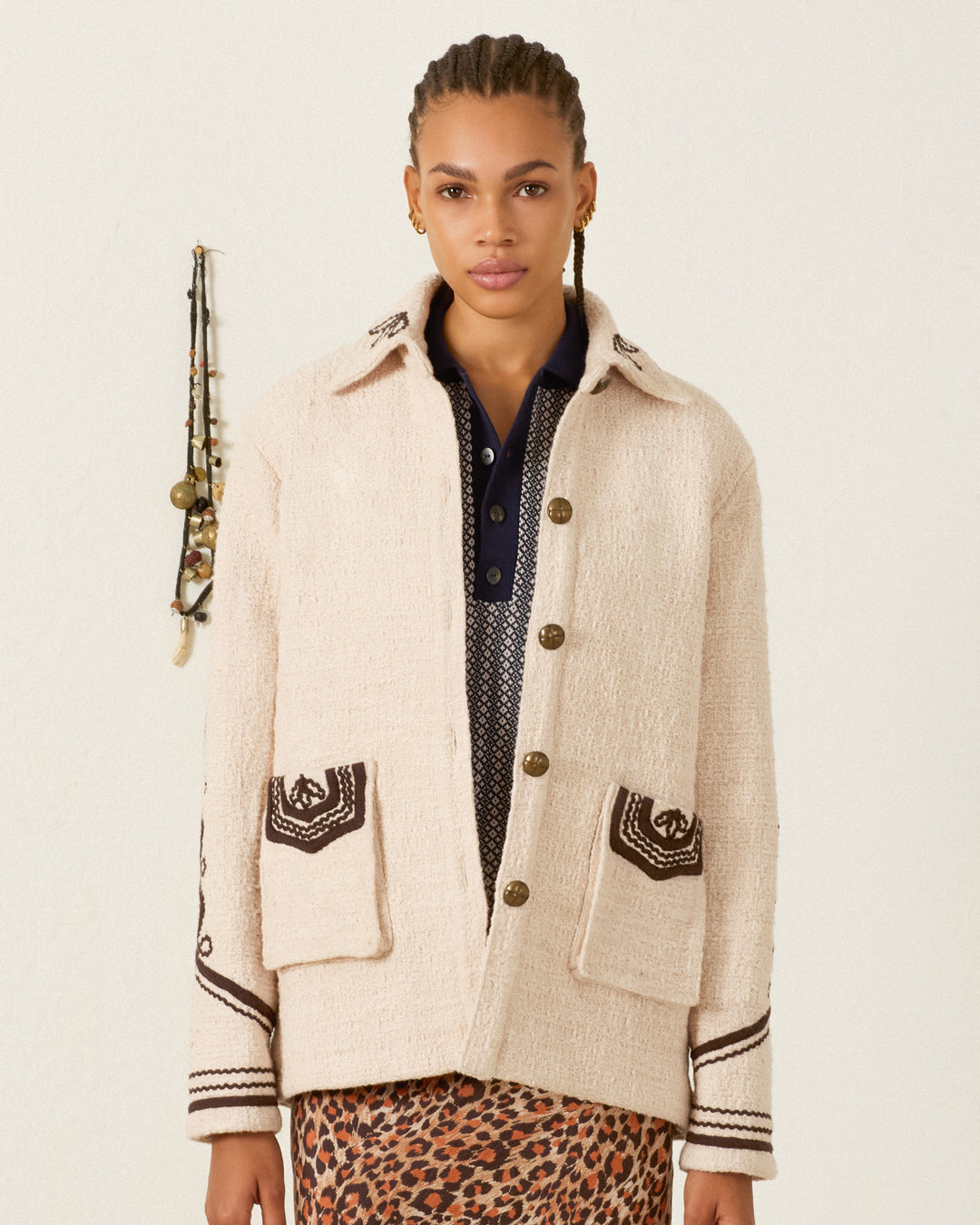 A person in the Jude Bushland Cord Jacket—a cream-colored piece with black patterns accenting the pockets and sleeves—stands against a light wall, necklaces hanging in the background. This vintage-inspired jacket showcases elegant soutache embroidery that lends an artistic touch to its sophisticated design.