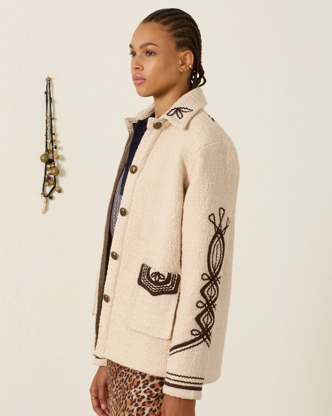 Person standing in profile wearing the cream-colored Jude Bushland Cord Jacket with cotton bouclé texture and black soutache embroidery, paired with an animal print skirt and braided hair. A small beaded wall hanging is visible in the background.
