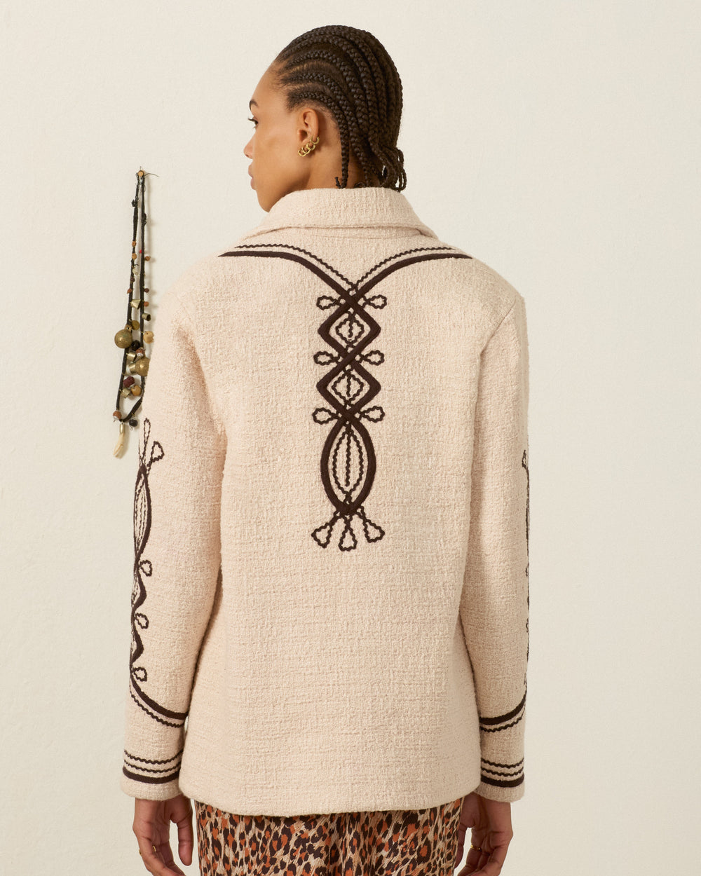 A person with braided hair wears the Jude Bushland Cord Jacket—a vintage-inspired cream cotton piece featuring black soutache embroidery in geometric patterns on the back and sleeves—while standing against a plain wall adorned with hanging jewelry.