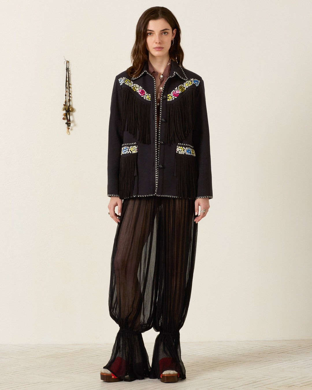 A woman stands against a plain wall, wearing the Jude Cross Bloom Jacket—a black fringed piece adorned with floral hand embroidery—paired with sheer black pants and red sandals. A set of hanging ornaments is visible on the wall beside her.