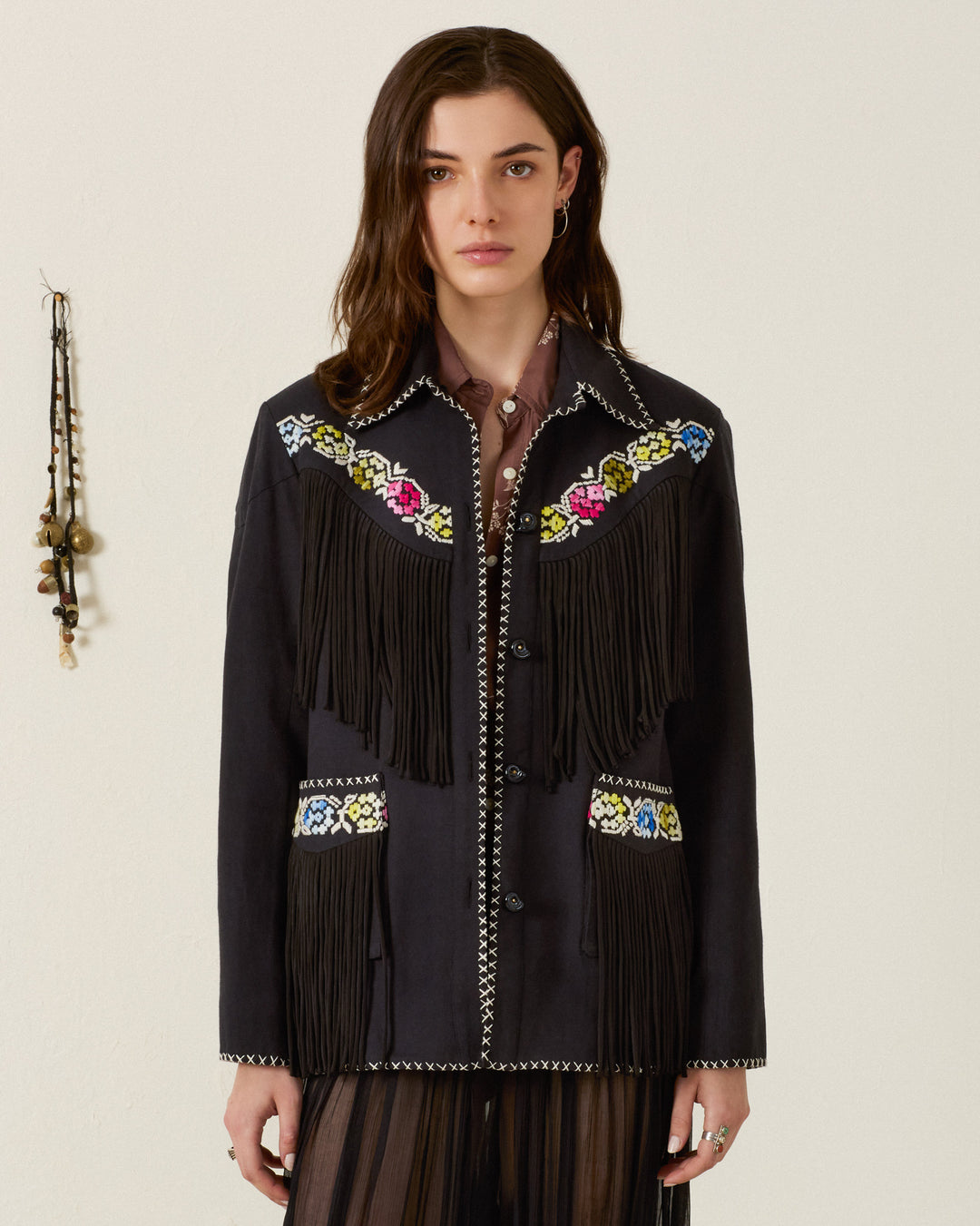 A woman wears the Jude Cross Bloom Jacket—a black, Western-style piece featuring floral hand embroidery, white cross-stitch detailing, and handcrafted nautilus shell buttons—over a brown collared shirt and a dark skirt.