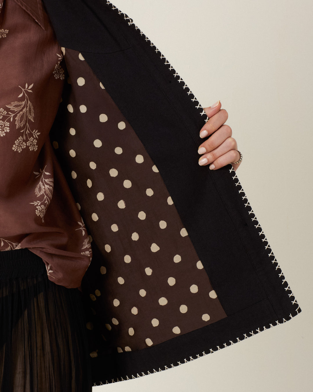 A person holds open the Jude Cross Bloom Jacket to reveal its brown lining with large white polka dots, accented by handcrafted nautilus shell buttons. The individual wears a brown floral-patterned top and a black pleated skirt.