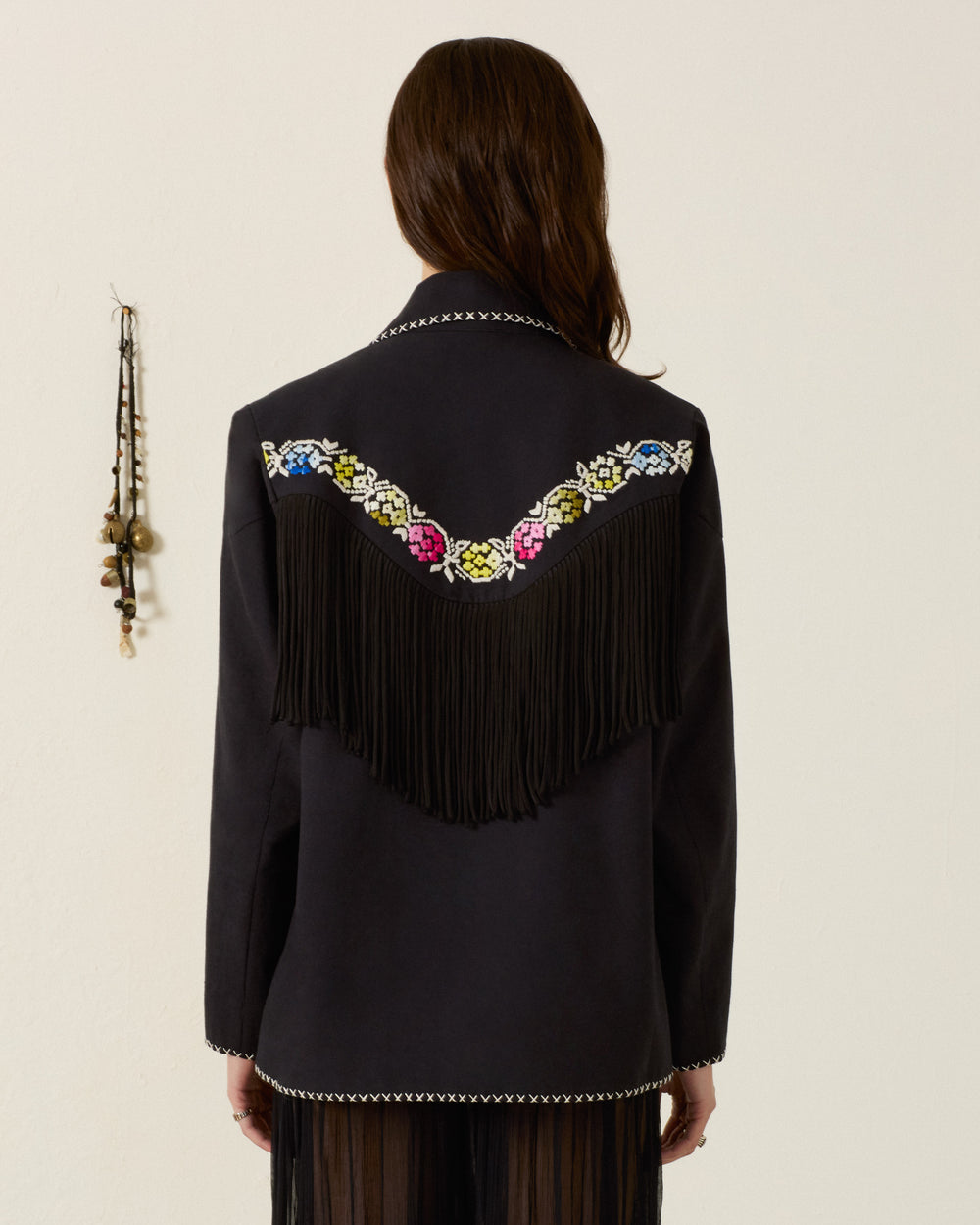 Wearing the Jude Cross Bloom Jacket—a black fringed piece adorned with beaded multicolor details and striking black fringe along the back—a person stands against a plain light wall with hanging ornaments to the side. Exquisite floral hand embroidery enhances its bold design, offering a unique artisanal touch.