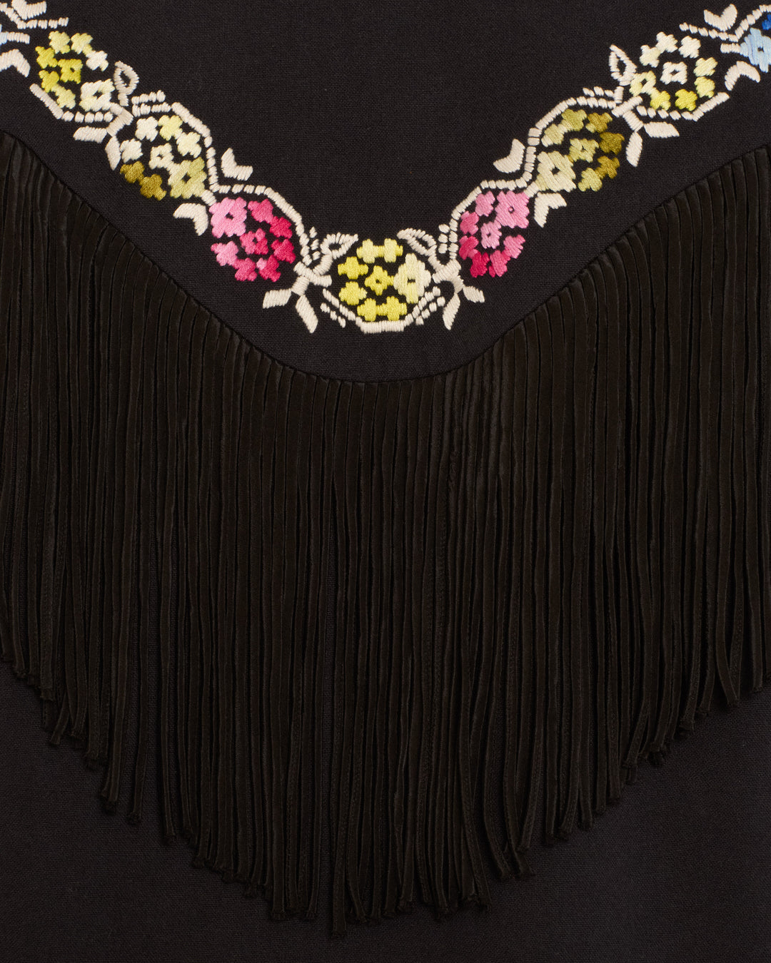 The Jude Cross Bloom Jacket features black fabric with a row of colorful floral hand embroidery along the top and long black fringe hanging beneath the embroidery.