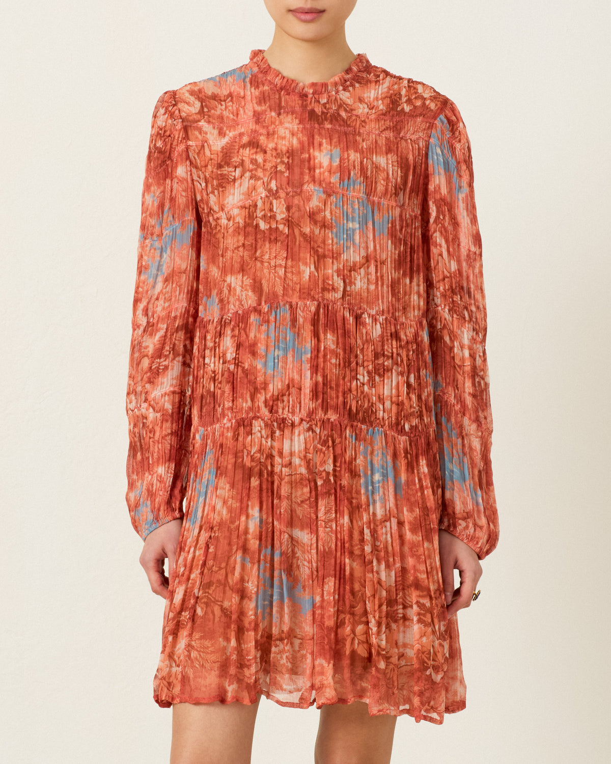 A person wearing the Juno Heritage Canopy Dress—a long-sleeved, knee-length dress adorned with a red, orange, and blue tie-dye pattern and featuring a delicate ruffle-trimmed neckline—stands against a plain light background.
