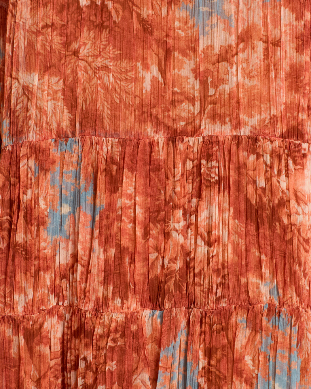 Close-up of pleated cupro-chiffon fabric from the Juno Heritage Canopy Dress, featuring a rust orange and light blue floral pattern inspired by a sylvan landscape, with gathered seams, textured vertical lines, and glimpses of a ruffle-trimmed neckline.