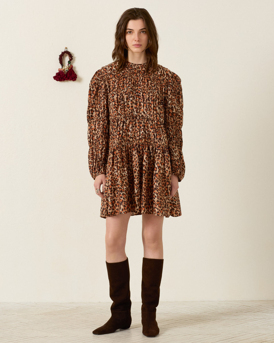 A woman stands indoors wearing the Juno Leopard Trek Silk Dress—hand-loomed in brown and orange patterns—with black knee-high boots; a small decorative item hangs on the wall behind her.