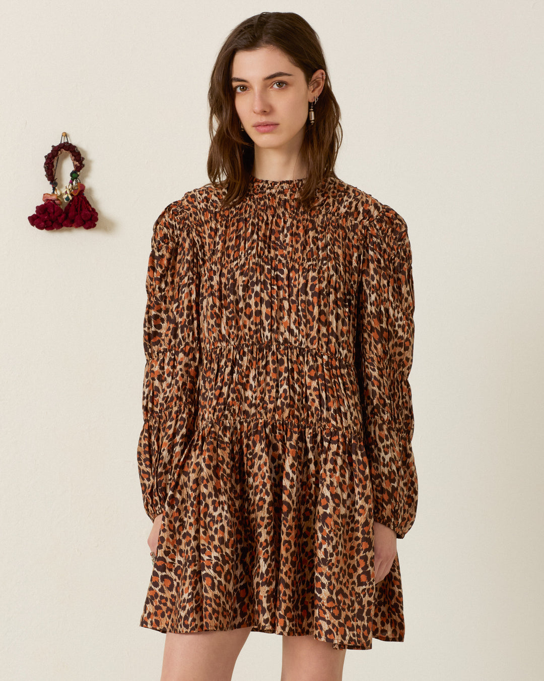 A woman stands indoors with a neutral expression, wearing the Juno Leopard Trek Silk Dress—a hand-loomed, long-sleeve leopard-print piece. To her left, a small red decoration adorns the plain light-colored wall.