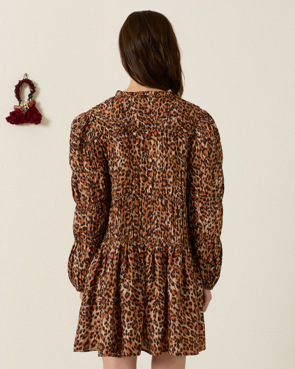 A person with long brown hair wearing the Juno Leopard Trek Silk Dress, a long-sleeved mini dress in a leopard-print pattern, stands facing a cream-colored wall. A red scrunchie hangs on the wall to the left.