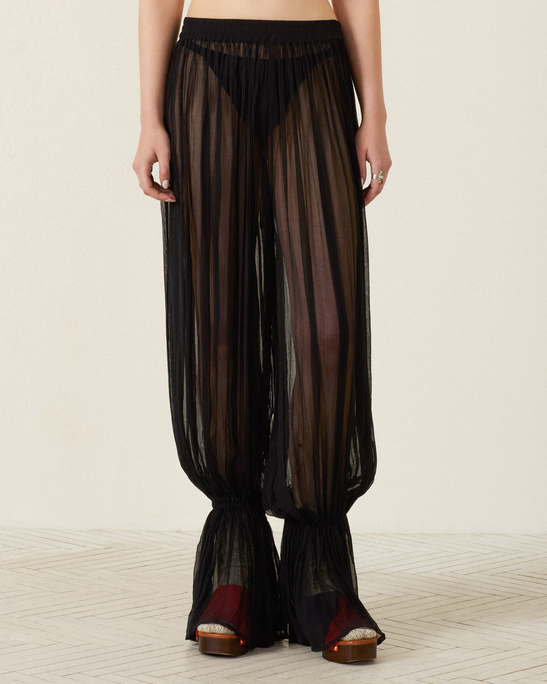 Wearing the Keats Onyx Chiffon Pant in sheer black pleated cupro-chiffon with flared hems and an elasticated waist, styled with visible black underwear and platform sandals, standing on a light-colored floor.