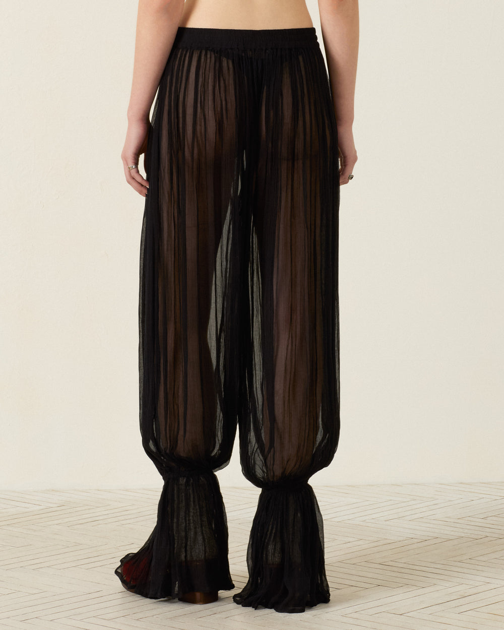 Wearing the Keats Onyx Chiffon Pant, featuring a sheer black wide-leg silhouette with gathered cuffs and an elasticated waist, this individual is seen from the back while standing on a light-colored patterned floor against a plain wall.