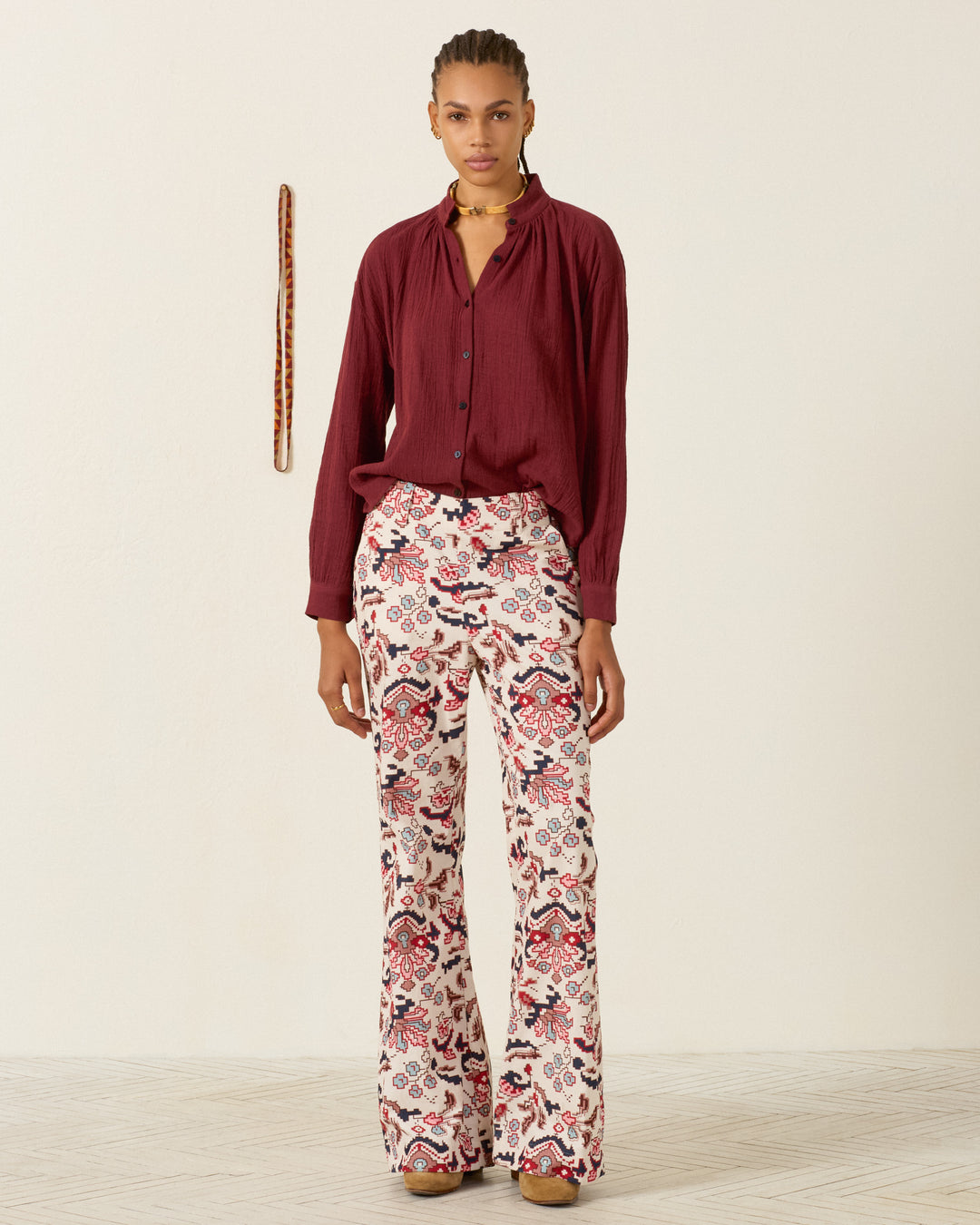 Person standing against a plain wall, wearing a long-sleeve maroon shirt, the Charlie Horizon Kilim Pant—high-waisted with a white and red kilim-inspired pattern—brown platform shoes, and a beaded necklace; a striped object hangs on the wall.