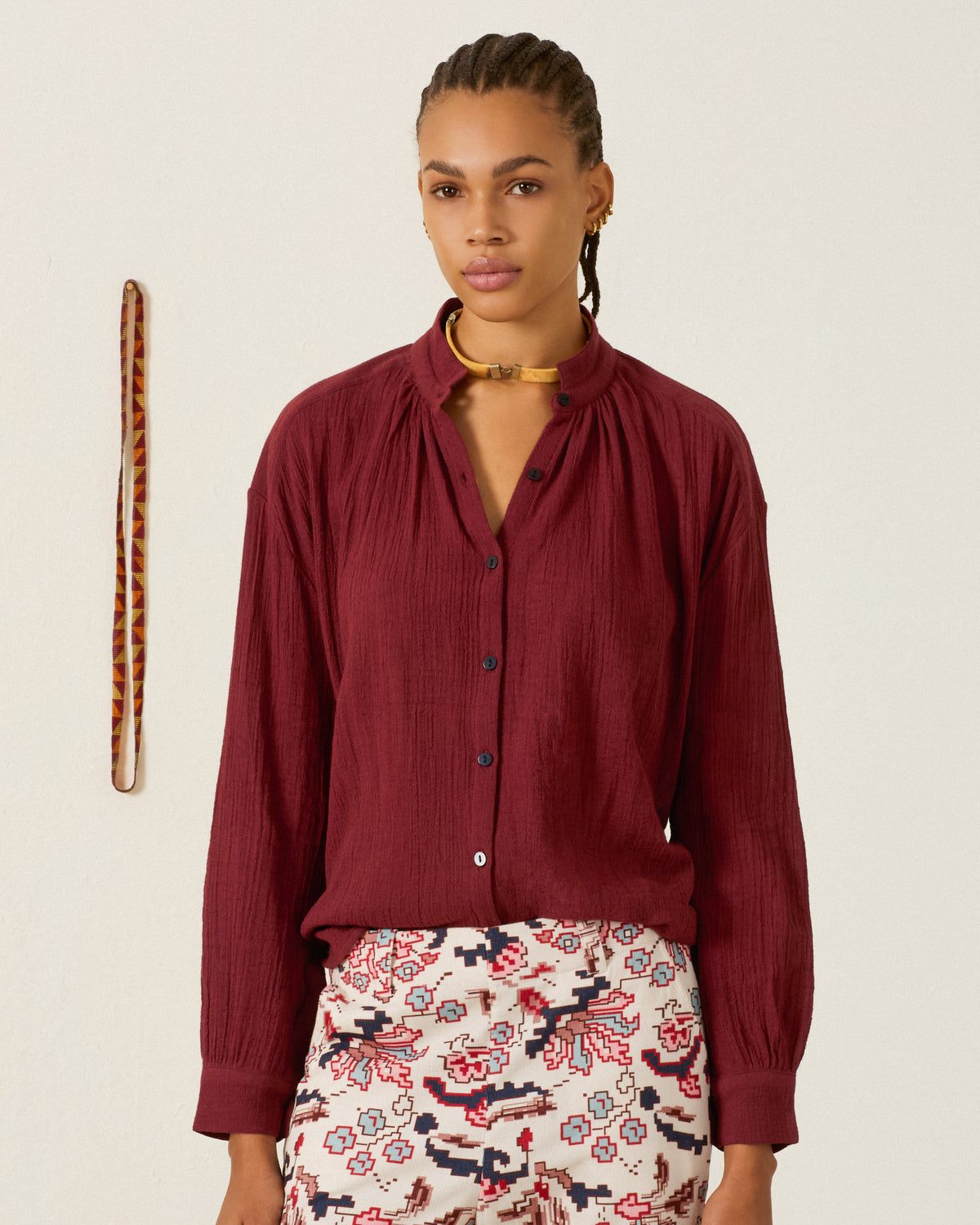 A person with braided hair wears the Kiki Mahogany Shirt, styled as a loose-fitting blouse with a patterned skirt, standing in front of a plain wall adorned with a decorative band.
