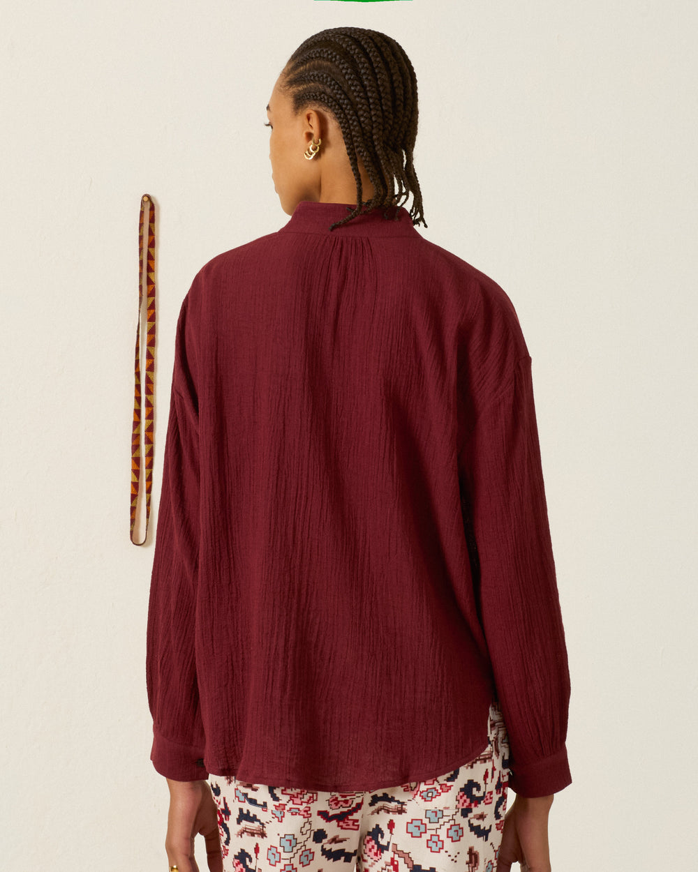 A person with braided hair, wearing the Kiki Mahogany Shirt and patterned pants, stands facing away from the camera. A patterned strap hangs on the white wall.