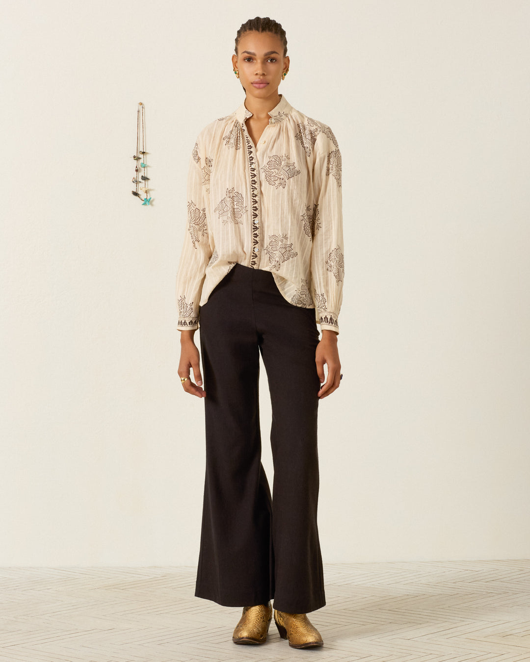 A person stands facing forward, wearing the Kiki Grapevine Stripe Shirt—a hand block printed cream blouse with an Edwardian silhouette—paired with black wide-leg pants and gold shoes, against a plain light background with a hanging decorative piece on the left.