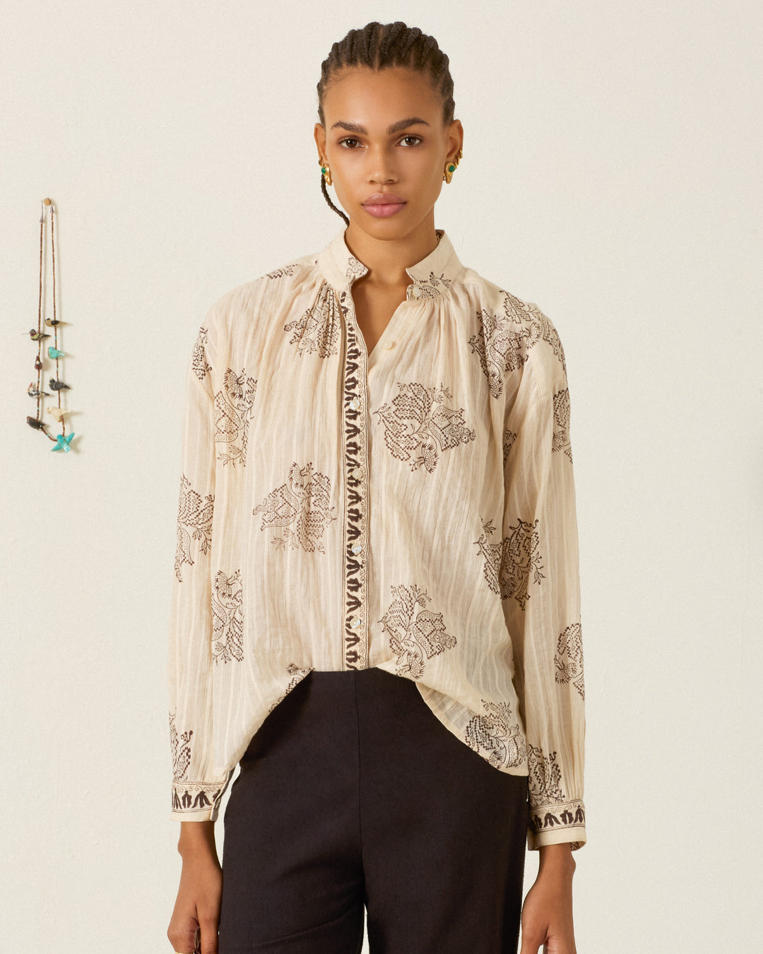 A woman stands in front of a light-colored wall, wearing the Kiki Grapevine Stripe Shirt—a beige, long-sleeve cotton dobby blouse adorned with dark floral patterns and subtle Edwardian silhouette details for a timeless look—paired with black pants. A decorative hanging is visible on the wall to her left.
