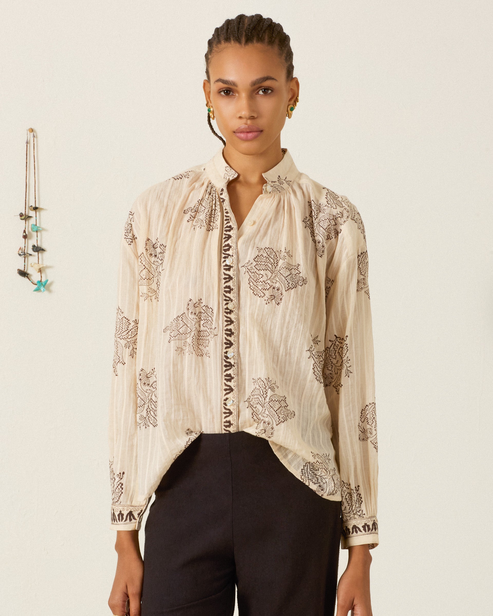 A woman stands in front of a light-colored wall, wearing the Kiki Grapevine Stripe Shirt—a beige, long-sleeve cotton dobby blouse adorned with dark floral patterns and subtle Edwardian silhouette details for a timeless look—paired with black pants. A decorative hanging is visible on the wall to her left.