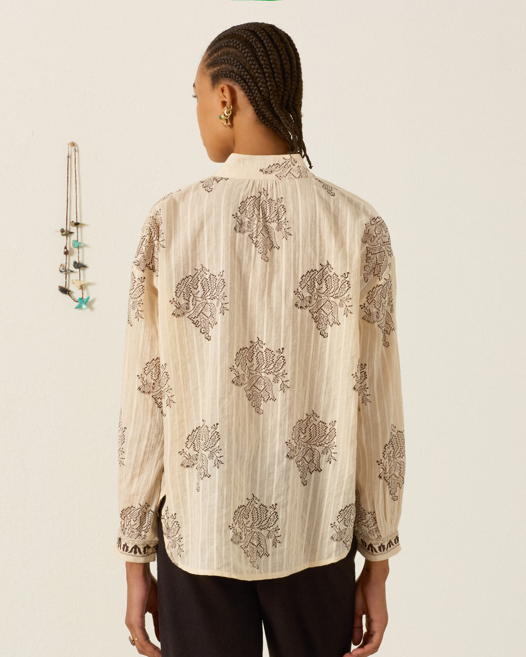 A person with braided hair, dressed in the Kiki Grapevine Stripe Shirt—a cream-colored, long-sleeved hand block printed cotton dobby shirt—and dark pants, stands facing away from the camera near a wall adorned with hanging necklaces.