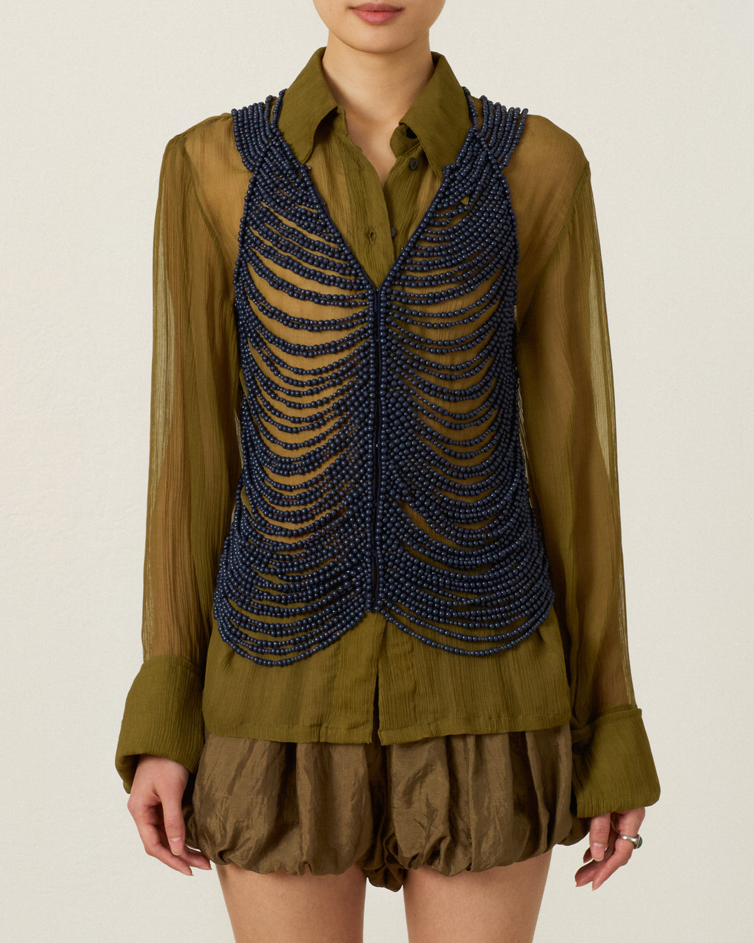 Wearing the Lania Indigo Wooden Top—a dark blue beaded vest arranged in horizontal rows—over an olive-green sheer blouse and brown textured shorts, accented with a handcrafted artisan body chain, the person stands against a plain background.
