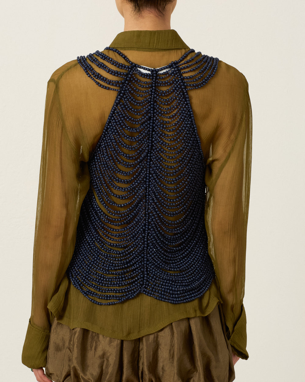 A person wearing the Lania Indigo Wooden Top, a sheer olive green blouse featuring an intricate blue beaded overlay reminiscent of a handcrafted body chain, seen from behind.