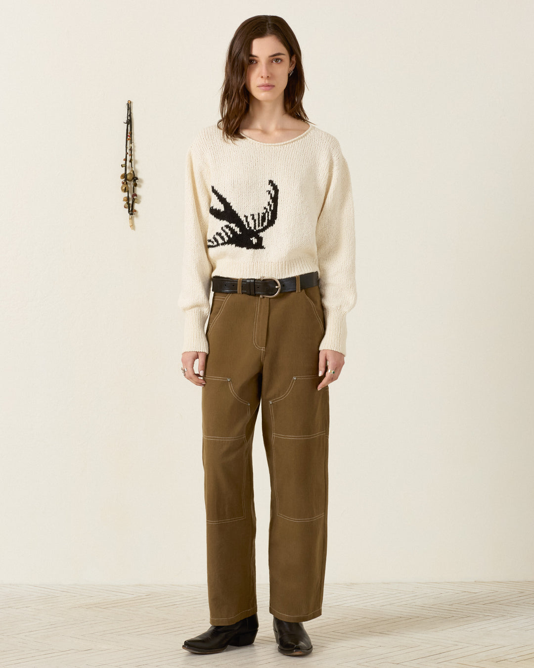 A person stands indoors wearing a cream sweater with a bird design, the Scottie Olive Branch Pant in brown high-waisted Khadi cotton, a black belt, and black shoes. A wall decoration with hanging objects is visible to the left.
