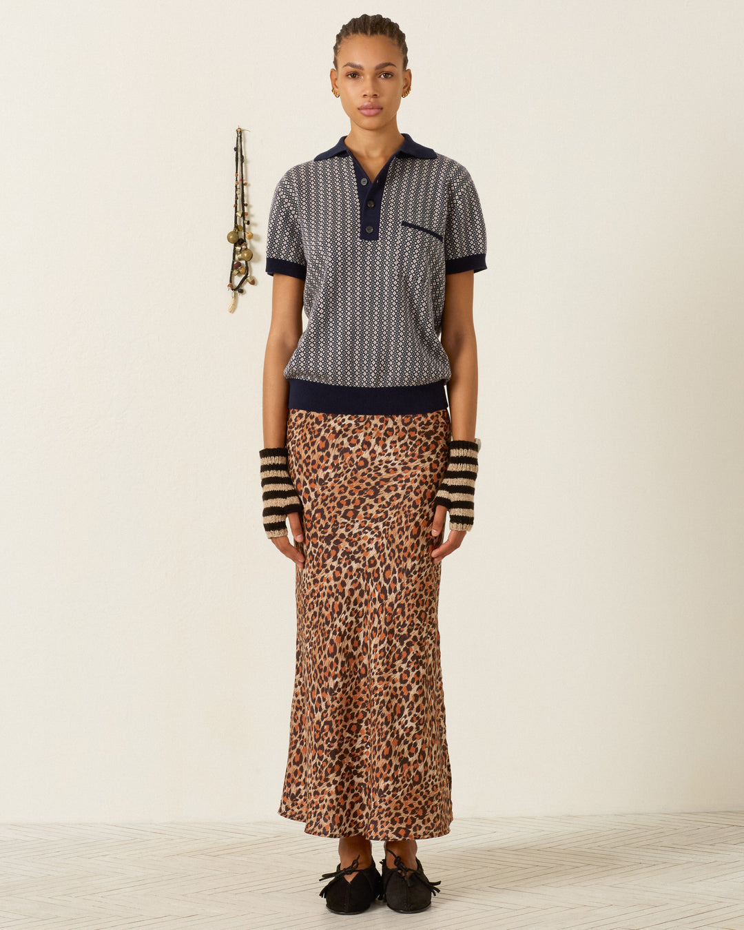 A person stands indoors wearing a patterned navy polo shirt, the Lyra Leopard Trek Silk Skirt featuring hand-loomed silk details, striped arm warmers, and black shoes. A necklace hangs on the wall behind them.