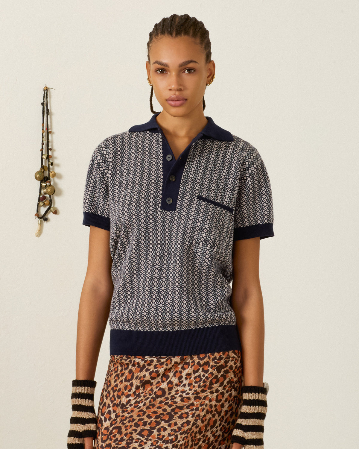 A person with braided hair wears the Lessi Cobblebud Knit Top, a vintage cropped polo featuring a geometric motif, paired with striped fingerless gloves and a leopard print skirt, standing against a plain wall with hanging decor on the left.