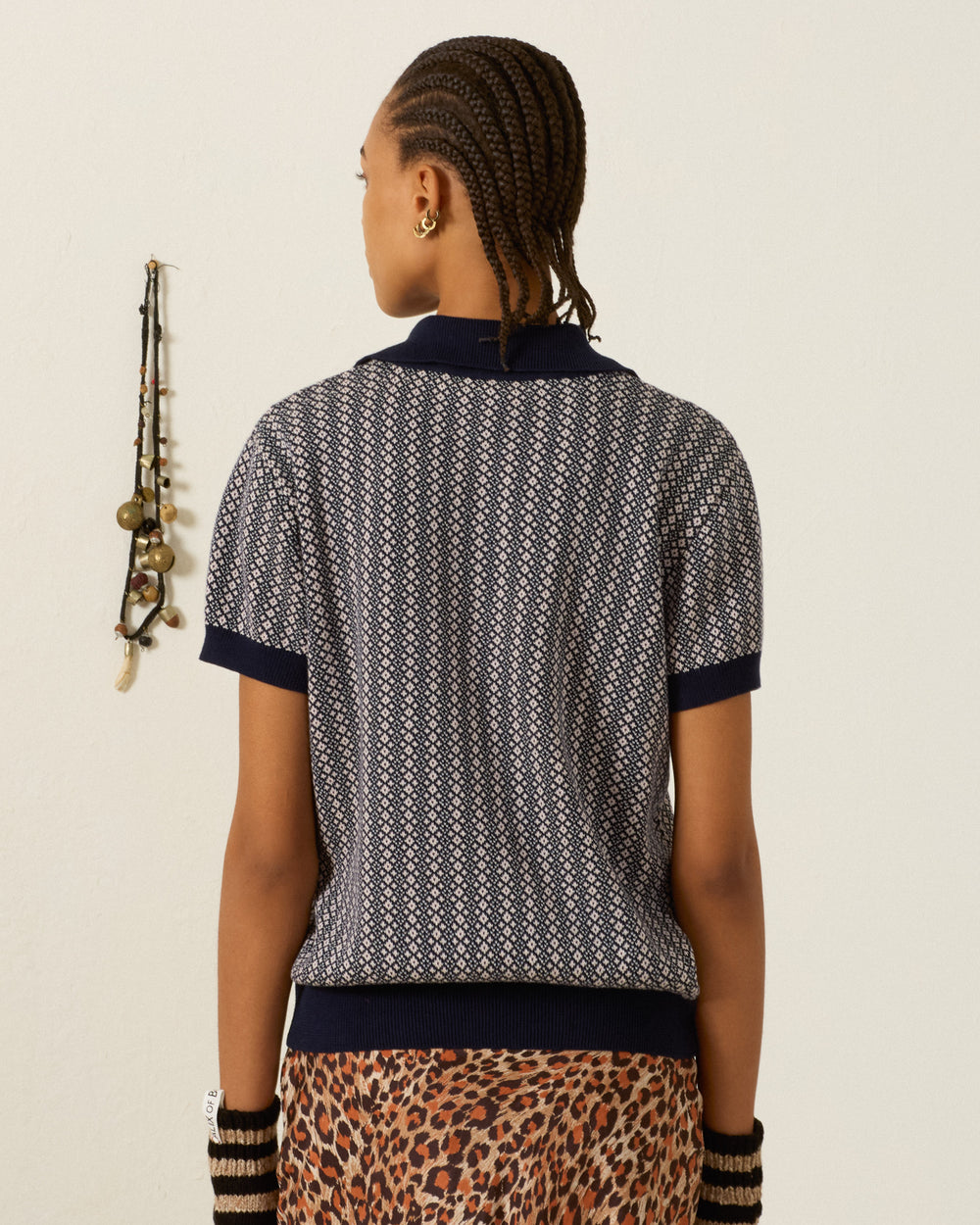 Person with braided hair, wearing the Lessi Cobblebud Knit Top—a vintage cropped polo featuring a geometric motif—and a leopard print skirt, stands facing away from the camera; jewelry hangs on the wall to the left.