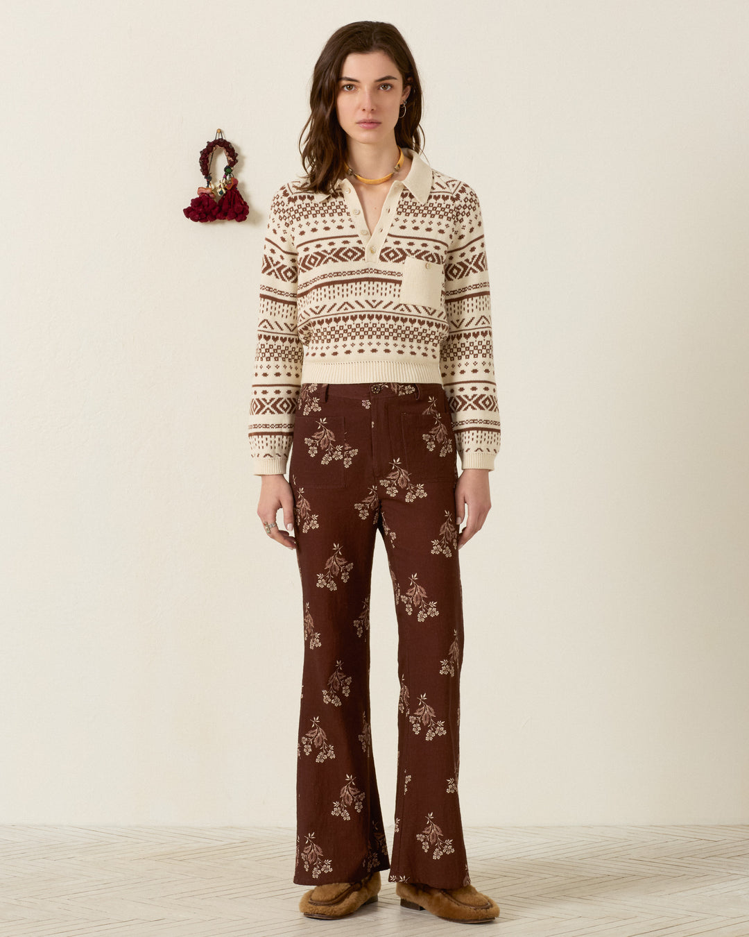 A woman stands indoors against a plain wall with a small decorative item, wearing a cream and brown patterned cotton sweater, tan shoes, and the Sonnet Doe Elderflower Jean in a brown floral flared-leg style.