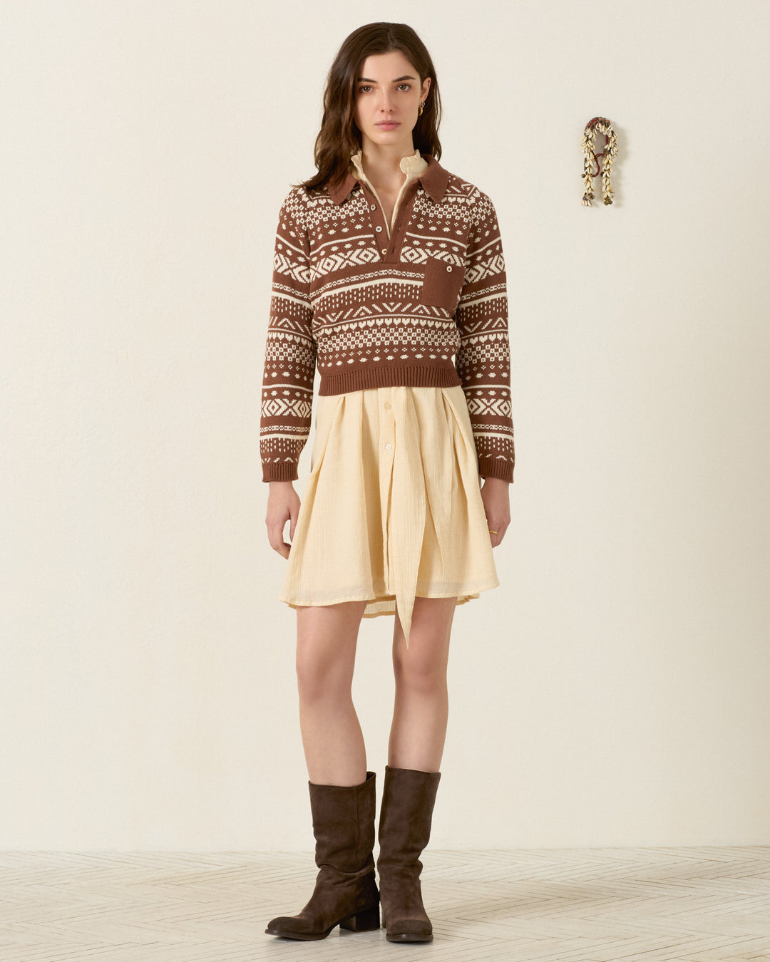 A woman stands indoors facing forward against a plain wall, wearing the Liza Mocha Fair Isle Top, paired with a cream-colored skirt and brown boots. A small decorative object is visible on the wall behind her.