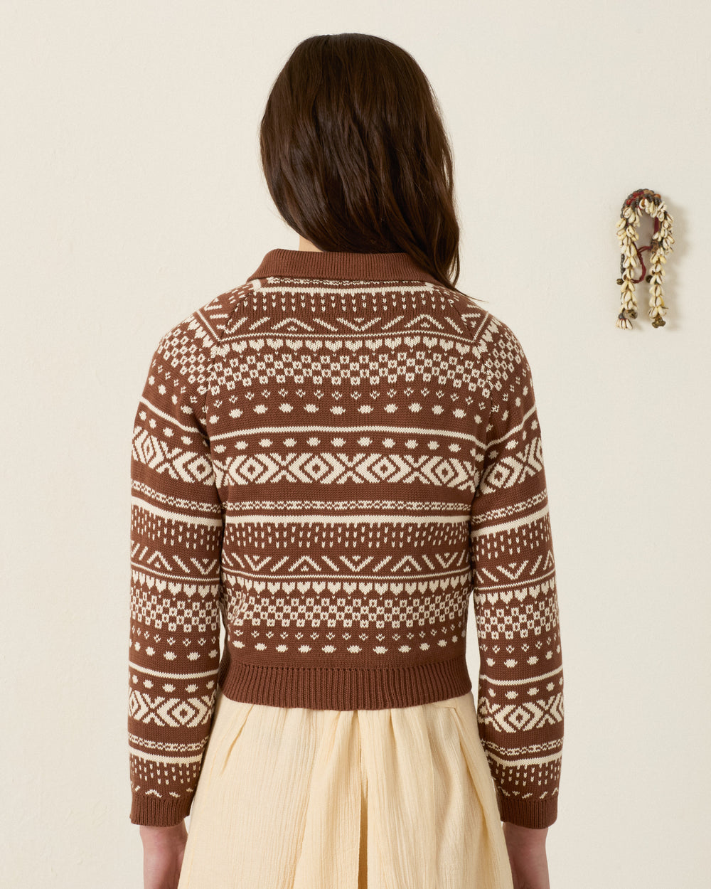 Wearing the Liza Mocha Fair Isle Top and a cream skirt, the person stands facing away against a plain wall with a small decorative hanging on the right.