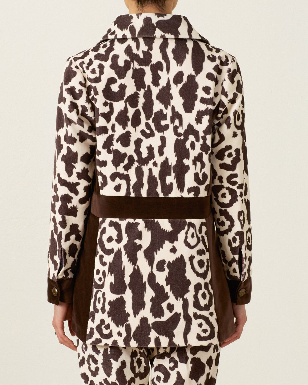 Wearing the Lydia Leopard Steppe Jacket, the person is seen from behind in a long-sleeve jacket and matching pants with a brown and white leopard print pattern, vintage silhouette, and solid brown bands at the waist and sides.