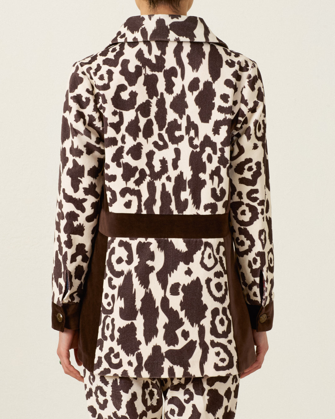 Wearing the Lydia Leopard Steppe Jacket, the person is seen from behind in a long-sleeve jacket and matching pants with a brown and white leopard print pattern, vintage silhouette, and solid brown bands at the waist and sides.